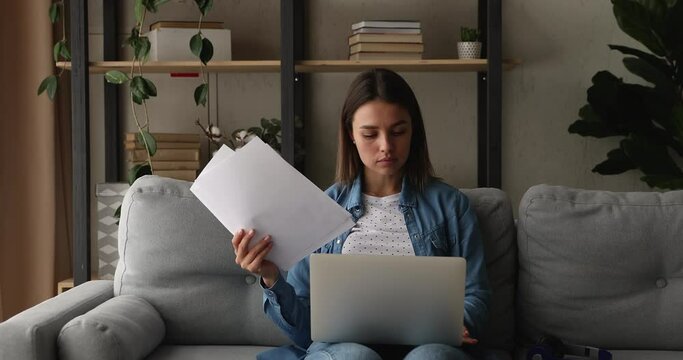 Young attractive serious woman sit on sofa put computer on laps holding papers do paperwork remotely, analyze documents data, learn and review contract terms, agreement conditions looking concentrated