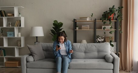 Young woman sit on sofa hold cellphone received message read sms stand up jumps looks crazy feel excited celebrate great news, got fantastic opportunity. Lottery winner enjoy moment of victory concept