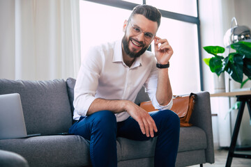 Fototapeta premium Confident stylish handsome bearded business man in glasses and formal smart wear is sitting on the couch in the modern office or room