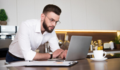Handsome confident businessman in smart casual wear is working with laptop at domestic kitchen