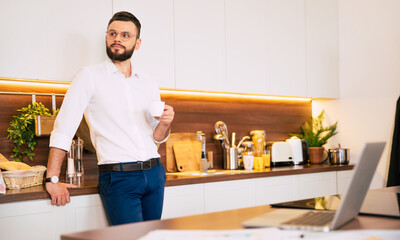 Fototapeta na wymiar Relaxed bearded confident businessman in smart casual wear drinks smells coffee and thoughtful at domestic kitchen