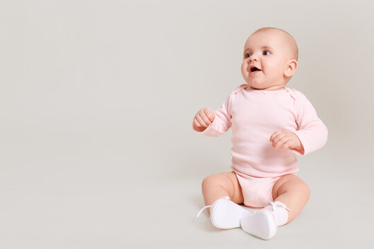 Happy excited infant child baby girl toddler wearing bodysuit and socks sitting on floor isolated on white background looking away with open mouth at free copy space.