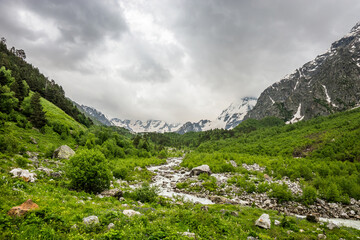 Summer cloudy landscape with Russian Caucasus snowy and woody mountains and small river at the foot of it..