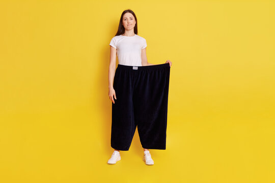 Full length photo of Young adult Caucasian girl wearing casual t shirt and too big size pants, looks at camera with puzzled expression and bewilderment, purses lips, isolated over yellow background.