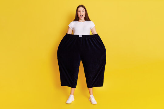Shocked astonished woman wearing old too big black trousers keeps hands in pants. looks at camera with open mouth and big eyes, has surprised facial expression, posing isolated over yellow background.
