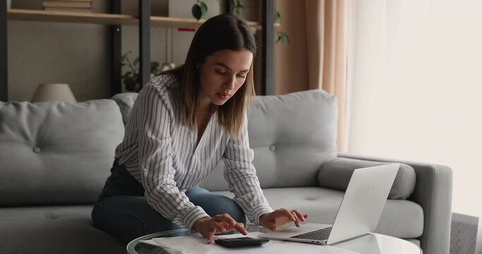 Young focused woman sit on couch at home calculates expenses on calculator, pay bills using on-line e-banking application on laptop looking concentrated. Managing personal budget, accountancy concept