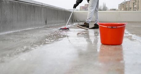 Man using broom and water to clean dirt from the rooftop. Red plastic bin on the right side of photography. He is wearing a black jacket, gloves, and gray sweat suit. Before and after situation.