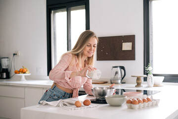 Cute blonde woman in pink blouse stays in kitchen, holds glass of flour, eggs are on table, going to make dough for cake. Person loves to cook. Girl prepares dessert according to delicious recipe