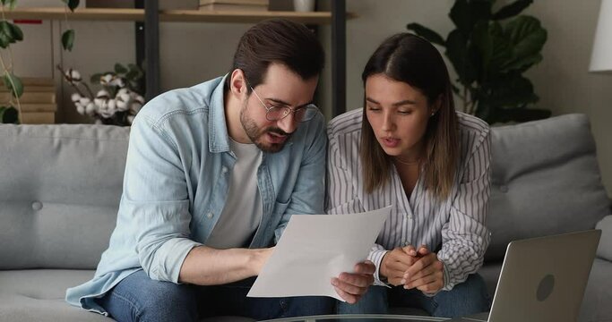 Serious millennial spouses sit on couch hold sheets read financial investment document, renters considering rental agreement. Married couple talking discussing terms, learn contract conditions concept