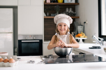 Small cute girl in special clothes and  funny chef cap stay on kitchen and make dough for cake. Making cookie at home on kitchen, kid get fun enjoy cook. Child make dessert