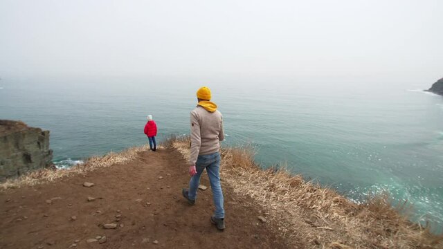 Father is filming his daughter who is standing on the cliff edge above the spring sea