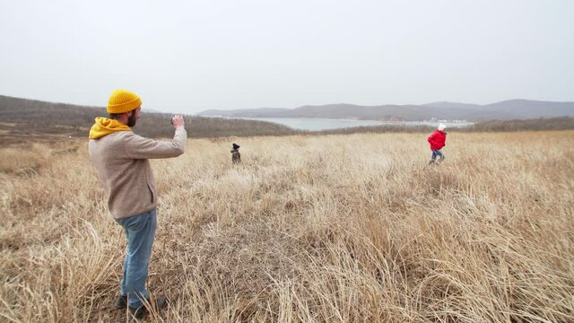 Father is filming his daughter and son running and having fun in high dry grass. Sea bay is on the backround