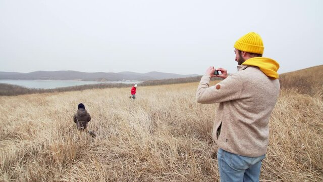 Father is filming his daughter and son running around and having fun in high dry grass. Sea bay is on the backround
