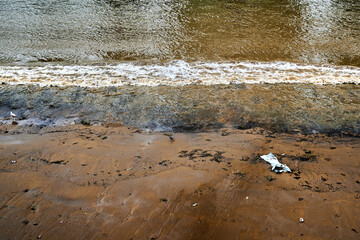 Looking down on river thames river bank exposed by low tide and a breaking wave and single piece of litter \ rubbish \ garbage