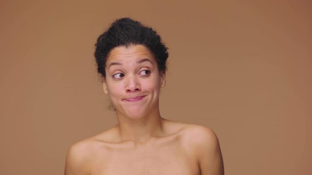 Beauty portrait of young African American woman saying oops, shrugging and confused looking away. Black female model posing on brown studio background. Slow motion ready, 4K at 59.94fps.