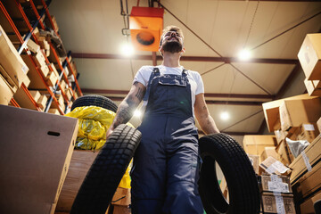 Low angle view of smiling hardworking blue collar worker in overalls carrying tires and walking in storage in import and export firm. © Dusan Petkovic