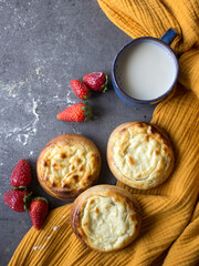 Authentic Russian open pies with cottage cheese, strawberry, ceramic cup with milk and bowl with cottage on a table. Yellow fabric background. Homemade pastries top view photo. 