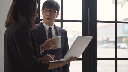 Business man and woman  in a good mood smile thinking with holding coffee cup, man and woman standing near the window talking to each other looking at the laptop.