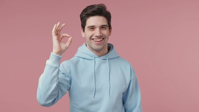 Handsome smiling cheerful brunet young man 20s wears blue streetwear hoodie say wow show okay ok zero fingers gesture isolated on pastel pink wall background studio portrait. People lifestyle concept