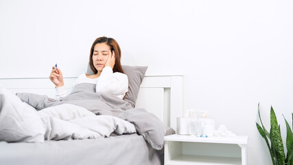 Sick Asian woman with flu virus lying in bed with high fever and measuring body temperature by thermometer and touching her forehead at her home.
