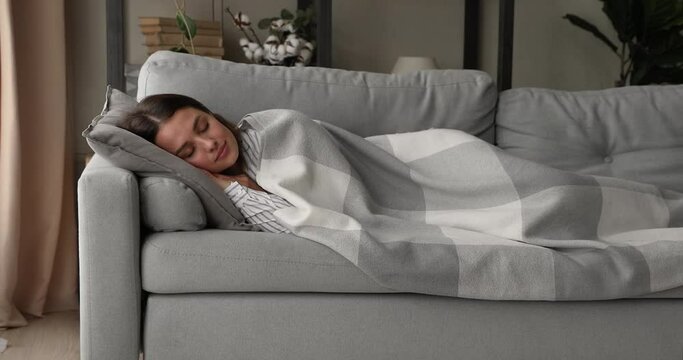 Young serene woman lying on sofa in light cozy living room enjoy fresh conditioned air and comfort sleep under warm plaid alone. Healthy day nap, repose and refreshment, lazy weekend and rest concept