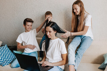 A young woman works at home with a laptop, together with the children, Children want to communicate with their mother, make noise and interfere with work. they play combing their mother's hair.