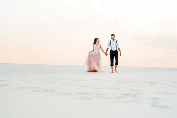 Fototapeta premium young couple a guy in black breeches and a girl in a pink dress are walking along the white sand