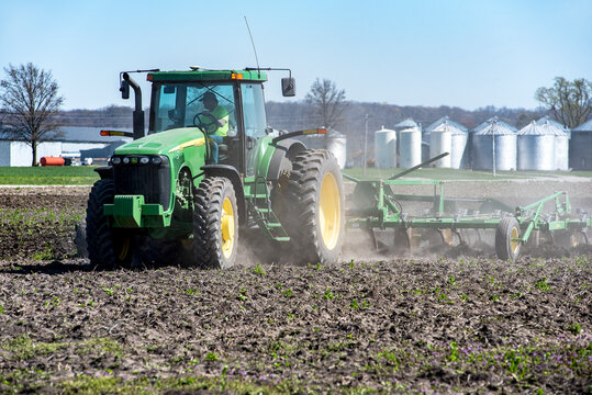 Illinois, USA, April 2, 2021 - Farmer in tractor plowing farm field preparing soil for spring corn planting with corn grain bins and silos in background.