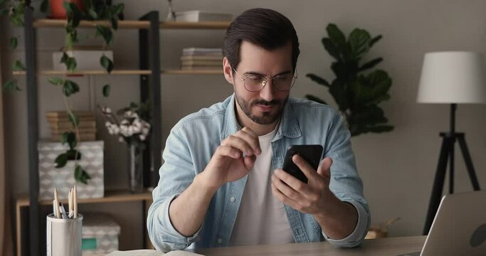 Millennial business man sitting at workplace desk holding modern smart phone using mobile business application, share text messages, communicates remotely, take break spend time on internet concept