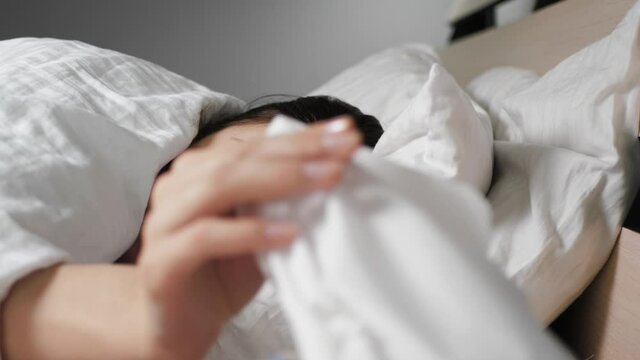Cold, flu, COVID-19 simptoms. Sick woman with cold and flu simptoms lies in bed under blanket and takes out paper napkin, blows her nose and wraps herself in blanket