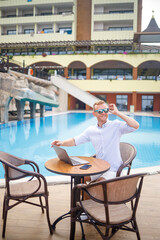 Fototapeta na wymiar Successful handsome male businessman in sunglasses works at a laptop sitting near the pool. Remote work. Freelancer