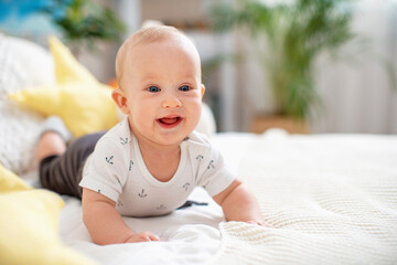 cute little baby is lying on bed on a white blanket at home and smiling. a small curious baby of 4 months, Caucasian, lies on the surface in a bright cozy room against the background of indoor plants