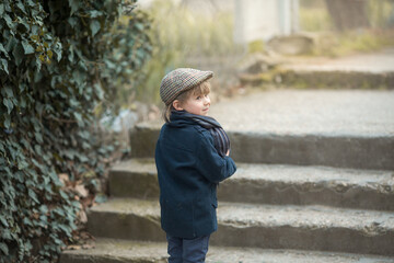 a beautiful little boy in a blue coat and scarf stands with his back to the street and turns around, hiding something under his coat. Portrait of a hipster or informal person.