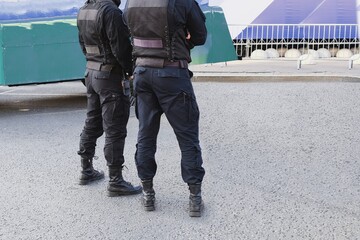 The back view of police officers wearing black uniform patrolling streets with walkie-talkie. Security guards in military outfit controlling city. Policemen trousers and boots of modern design.