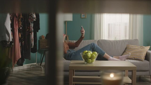 Beautiful girl in blue jeans and brown sweater lies on beige sofa on background of window, biting a red apple and and taking a selfie by phone. General shot. The camera moves to the right
