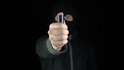 A man in a balaclava mask stands with a pepper spray. A bandit holds out a pepper spray to the camera. On a black background.