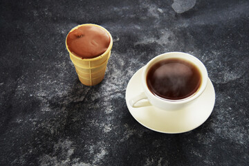 Ice cream chocolate in waffle cup with white ceramic mug of hot tea on dark concrete background