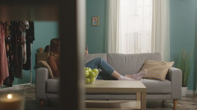 Young girl in blue jeans and brown sweater lies on beige sofa on background of window, biting a red apple and and speaks via video link.