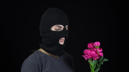 A Woman in a balaclava mask is standing with flowers. Bandit sniffs a bouquet of pink flowers on a black background.
