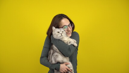 Obraz premium A young woman holds a cat in her arms and hugs him on a yellow background. Woman with glasses and a green sweater.