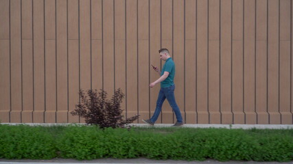 A young man goes with a phone in his hand. A man walks near the building and carries on correspondence in a smartphone.