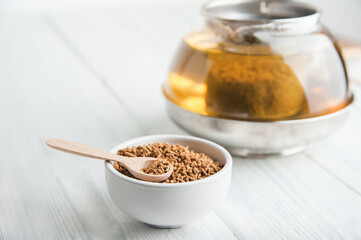 uncooked buckwheat in white plate and wooden scoop, on a white background close-up. Buckwheat tea and granules. Tea in transparent teapot. Space for text.
