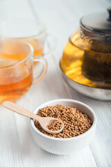 uncooked buckwheat in white plate and wooden scoop, on a white background close-up. Buckwheat tea and granules. Tea in transparent teapot. Space for text.
