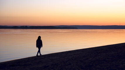 A young woman walks along the beach along the river at sunrise. Silhouette of a girl. Goes to meet the dawn.