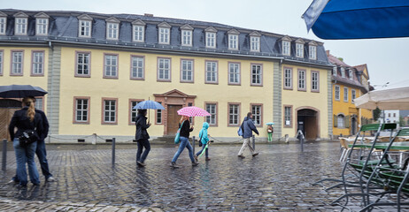 People walk past Goethe's house with umbrellas. Weimar. Am Frauenplan. Germany. The house is blurred, the people sharp. What would Gothe write today?