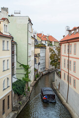 Fototapeta na wymiar Narrow canal between the ancient architecture of the old town in Prague. A tourist boat with the flag of the Czech Republic is sailing along the river. Houses in greenery, with terraces, balconies