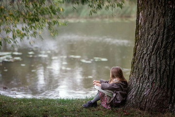 Girl reading book by the lake in the park. Green lake and child by the tree.