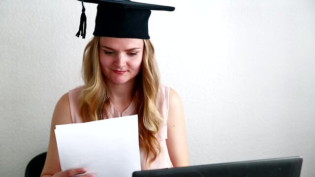 Woman student in masters hat takes an online exam, reading a diploma behind a laptop. Online education, graduation