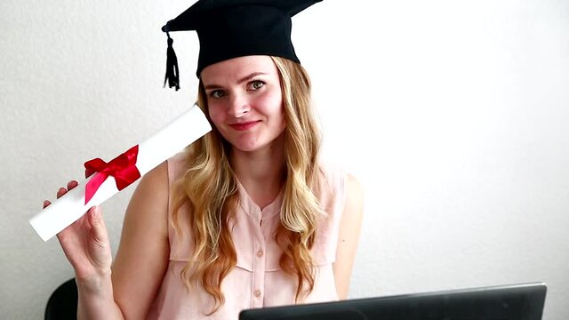 Happy woman student in masters after online exam, dancing with diploma behind a laptop. Online education, graduation concept