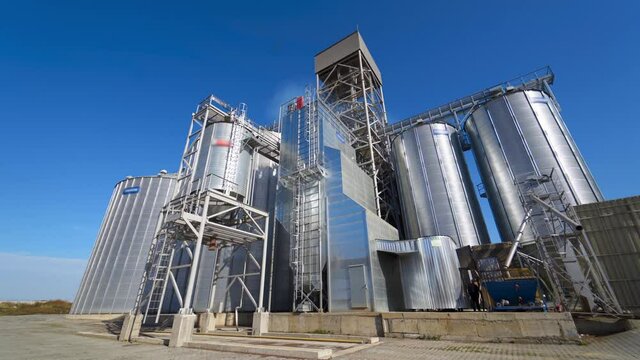 Modern Industrial Plant. Exterior Of A Large Agricultural Complex With Silver Grain Elevators For Storage Crop. Innovative Manufacture In Sunny Day. Front View.